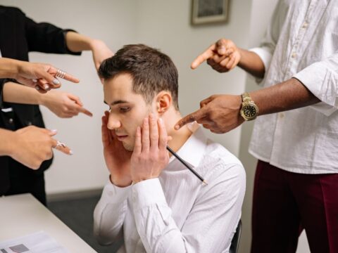 A stressed man at the office surrounded by pointing fingers, representing workplace bullying and stress.
